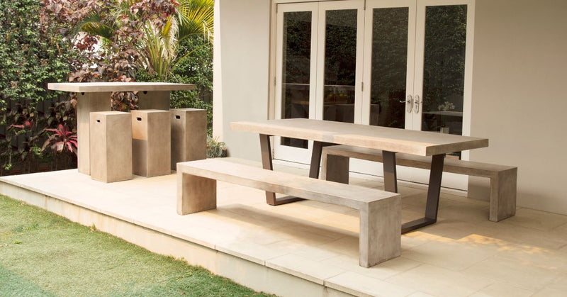 A Zen Outdoor Dining Setting with a concrete table and benches, and a matching Zen Outdoor Bar Setting with stools, arranged on a patio beside a house with sliding glass doors, overlooking a garden.