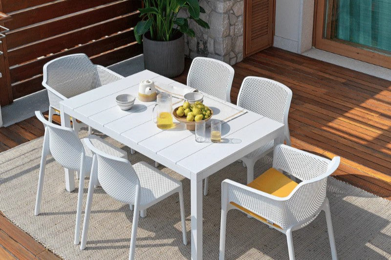A white outdoor resin dining table surrounded by six matching white resin dining chairs, creating a complete set. The arrangement is on an outdoor wooden deck with a rug underneath. The table is set with fruit & tea, with a pot plant in the background.