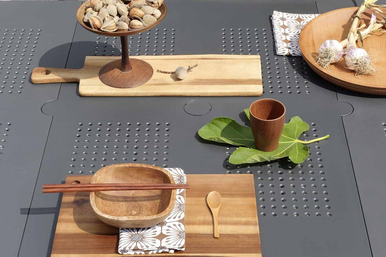 Close-up of an outdoor dining setting featuring a resin extension dining table and three outdoor resin armless dining chairs. The table is set with fancy wooden cutlery and adorned with plants and food, all situated in an outdoor area.