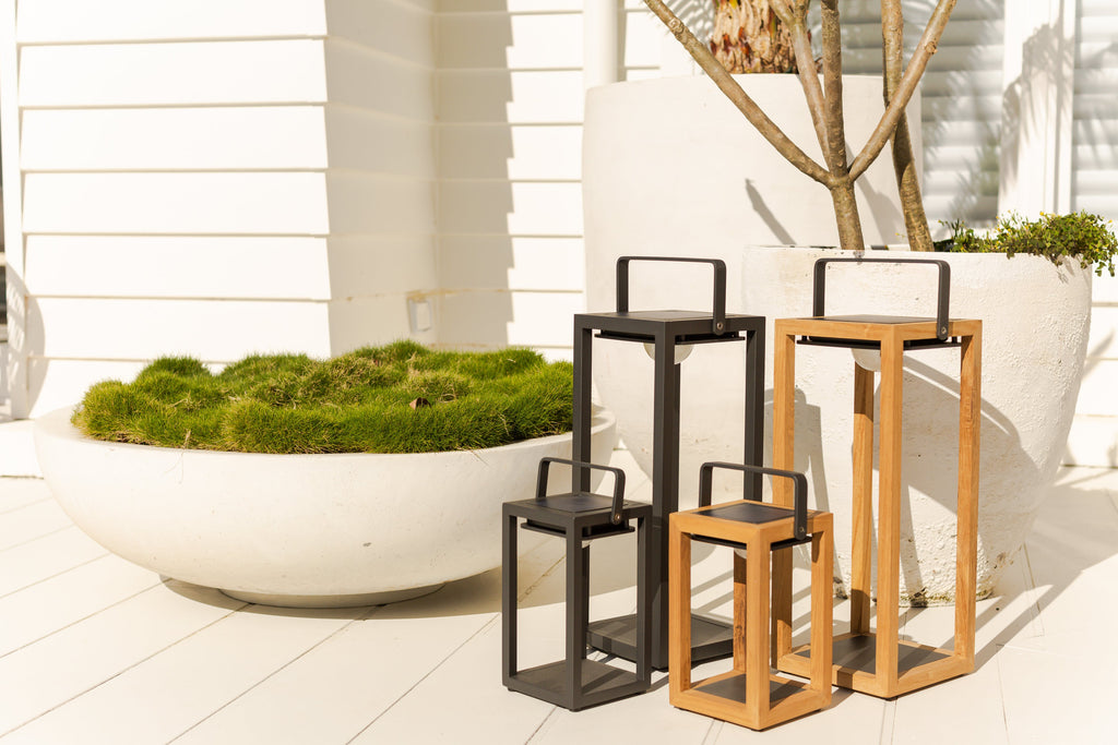 Lincoln Solar Lamps in different sizes with sleek aluminium and teak frames, showcased on a sunlit white tiled patio. Adjacent, a white planter holds vibrant greenery.