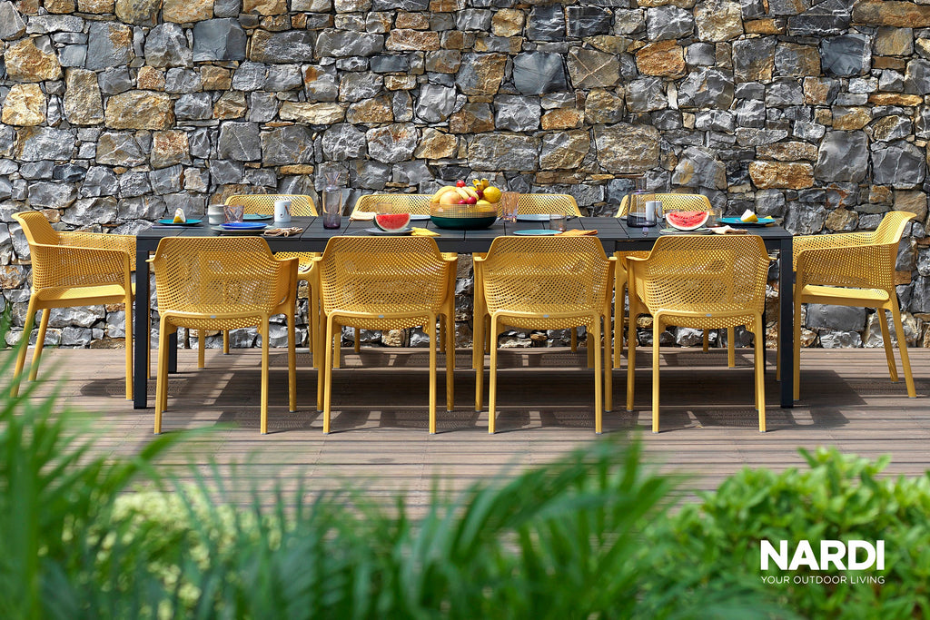 Pair of Nardi Doga Outdoor Resin Dining Armchairs in Marsala positioned on either side of a square black table, adorned with a decorative vase. The ensemble is set against a textured concrete wall featuring surrounding greenery and captivating shadows.