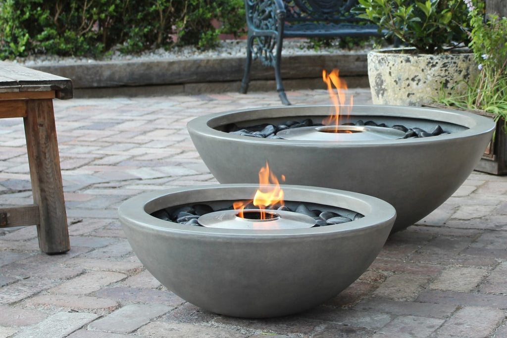 An EcoSmart Mix850 fire pit in bone color is the centerpiece in a cozy outdoor nook with built-in wooden bench seating, soft cushions, a rocking chair, and surrounded by lush tropical plants and ambient lighting.