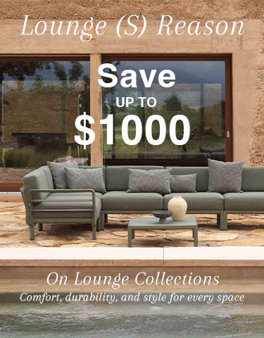 A modern grey outdoor lounge with 3 decorative pillows and an outdoor coffee table, set against a green wall. We see 2 pot plants & a cup of tea on the coffee table.