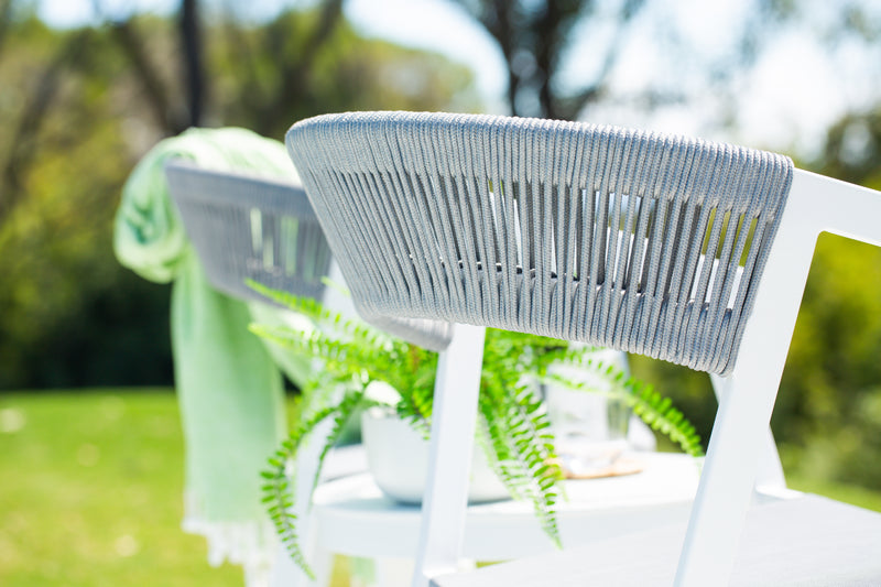 White Auto Rope dining chairs with grey rope backrests are set outdoors, one draped with a green throw and another holding a tray with a cup, against a lush garden backdrop.