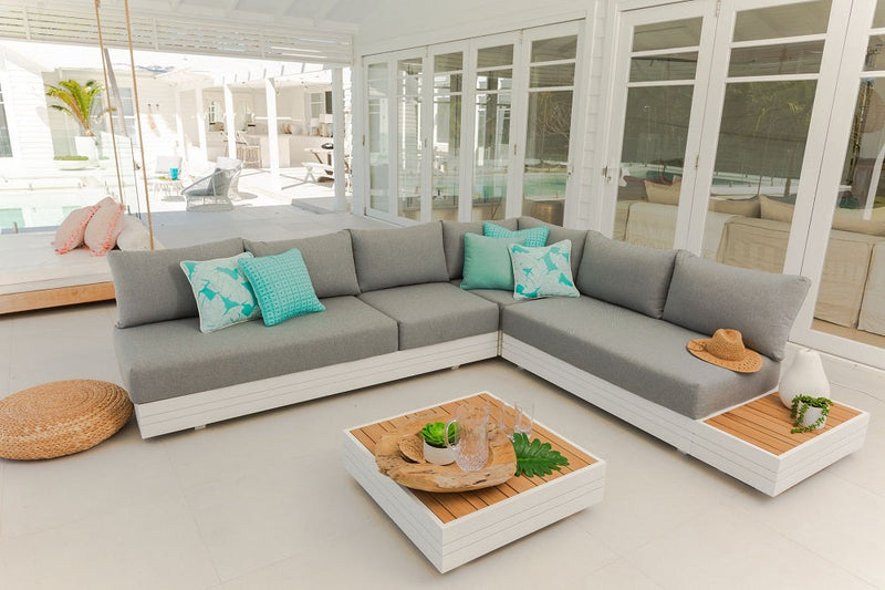 Hannover 6-Seater Outdoor Aluminium Modular Lounge with Coffee Table in White showcasing an elegant L-shaped gray sofa adorned with turquoise accent pillows, set against a modern, glass-enclosed patio with a matching white coffee table at the center.