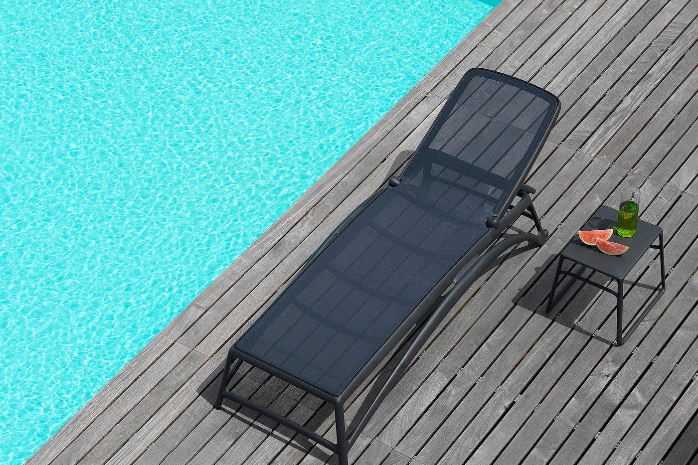 Black outdoor daybed on a wooden deck beside a pool. This daybed can also function as a sun lounge. There is a small coffee table beside the lounger, which has watermelon and a drink on it.