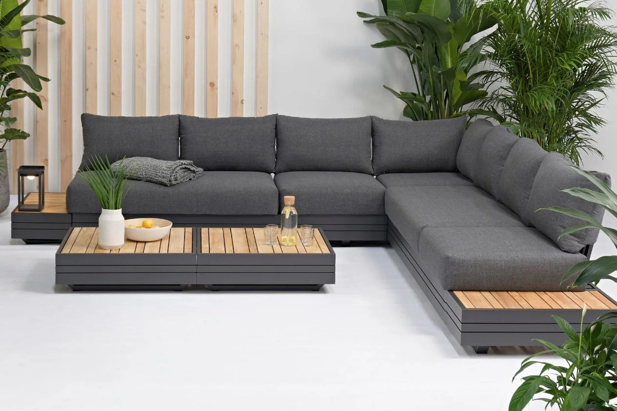Stylish aluminium & timber outdoor modular lounge with dark grey upholstery. The lounge is accompanied by a matching coffee table adorned with fruit and water, complemented by lush greenery in the background.