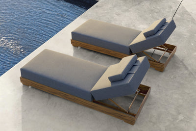 Two sun lounges with grey cushioning and wooden frames placed on cement by the seaside, bathed in relaxing sunset light.