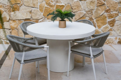A white 4 seater outdoor table with 4 matching chairs set against a textured stone wall, offering a charming outdoor ambiance.