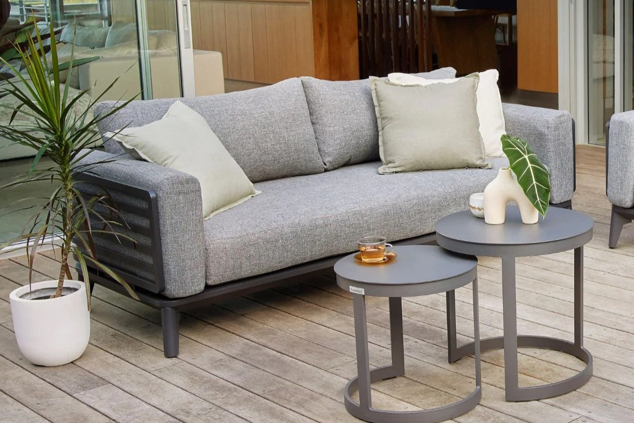 A modern grey outdoor sofa and two outdoor coffee tables on a wooden deck. Behind the sofa, the living room of the modern house is visible. Next to the sofa is a potted plant, and on the coffee tables are tea and a smaller potted plant.