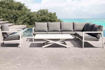 A white aluminium outdoor modular lounge set on a patio, accompanied by a white aluminium outdoor coffee table. Behind it, there's a stunningly blue body of water and some trees, with beautiful weather that resembles paradise.