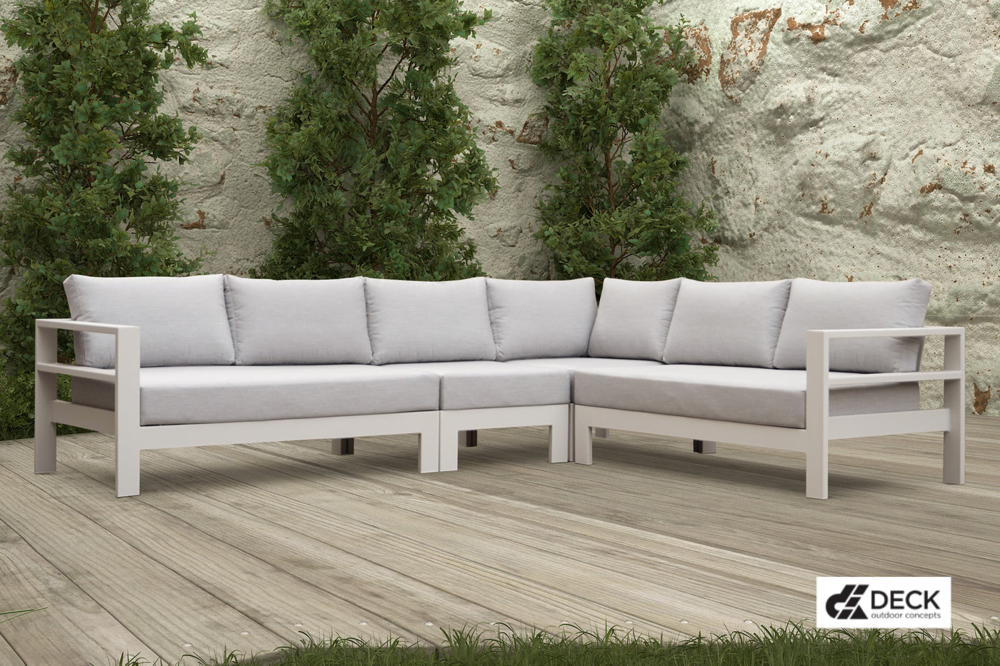 Deck Outdoor Concepts 6-seater Albury modular outdoor lounge in white, placed on a wooden deck, with a glimpse of greenery in the foreground, and a sandstone wall and three tall trees in the background.