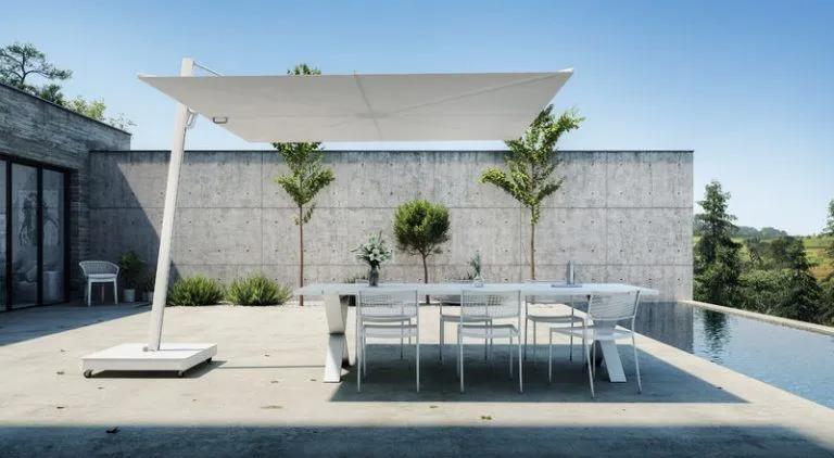 The Versa UX cantilever umbrella in white, providing shade over a modern outdoor dining area with white chairs, adjacent to an infinity pool, with a backdrop of a concrete wall and green landscape.
