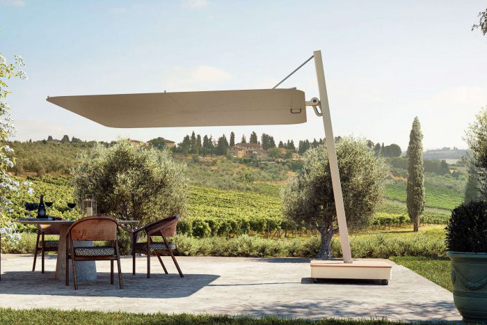 The Versa UX cantilever umbrella in white, providing shade over a modern outdoor dining area with white chairs, adjacent to an infinity pool, with a backdrop of a concrete wall and green landscape.