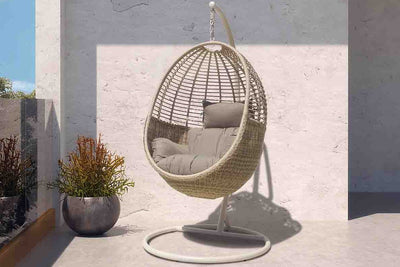 A stylish, modern hanging chair made of woven materials in a light neutral colour, suspended from a sturdy white metal frame. The chair is set in an outdoor patio area with greenery in the background & features some light grey cloud-like cushions.