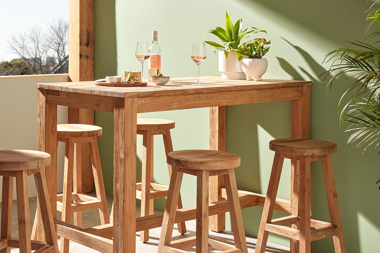 Outdoor bar table made of wood, surrounded by five matching wooden bar stools, set against a green wall in an outdoor area. The table has cheese, wine, and pot plants on it.