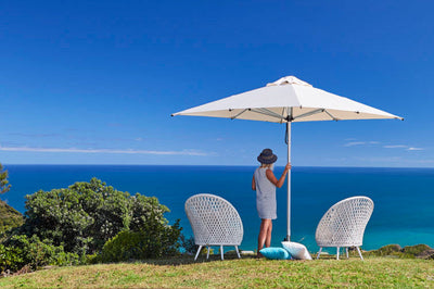 White garden umbrella providing shade for a woman standing on a hillside, with two outdoor chairs, enjoying a seaside view in the distance.