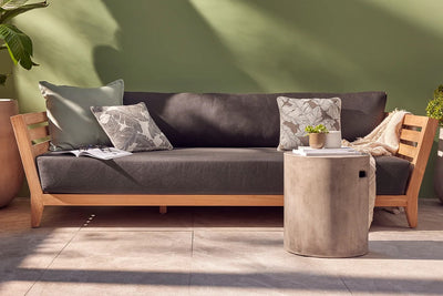 Timber outdoor lounge with dark charcoal grey cushions and three throw pillows, set against a green wall. A concrete coffee table in front holds two small pot plants. Two larger potted plants are flanking the lounge (on each side).