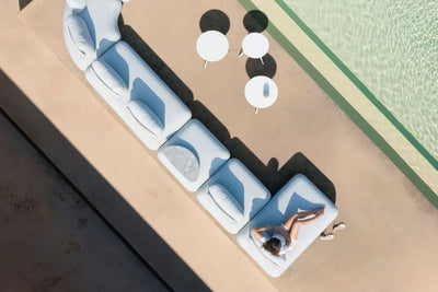 Bird's eye view of a woman relaxing on a white 6+ seater outdoor modular lounge by a pool on a sunny day. Three small outdoor coffee tables accompany the lounge.