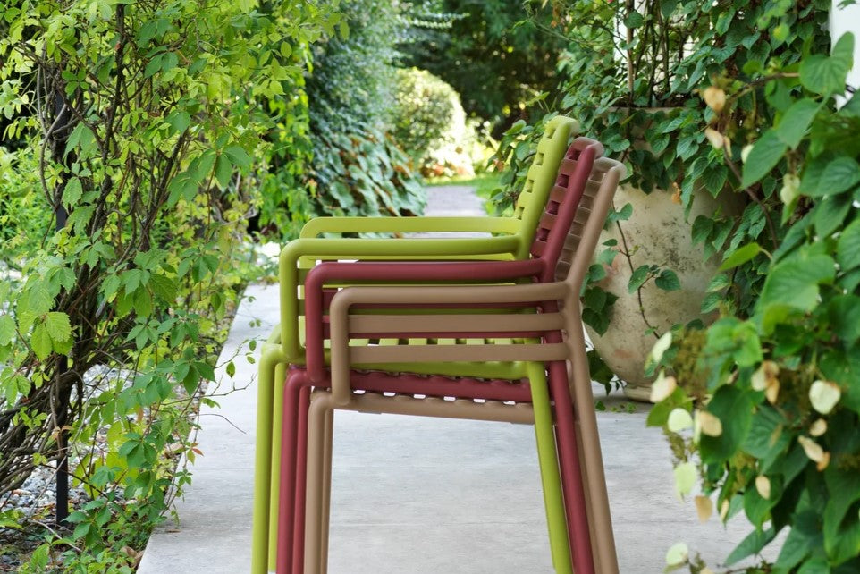 A stack of 3 colourful resin outdoor chairs positioned on a stone walkway, surrounded by natural elements, creating a serene outdoor setting.
