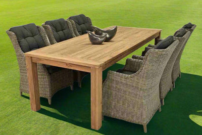 A wooden outdoor table surrounded by six outdoor wicker chairs, making a complete outdoor wicker dining set, all set on a big grassy field.