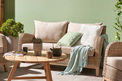 Outdoor wicker lounge with a coffee table against a green wall. The table holds tea, a pot plant, and macarons on a plate. A blanket is draped over the lounge. A vine plant is visible on the right, and a potted plant is in the background on the left side.