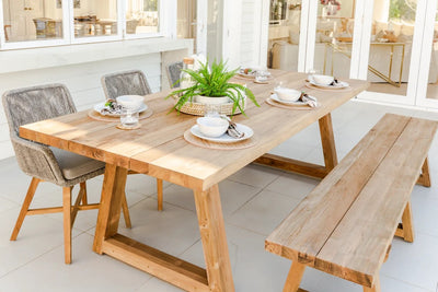 An outdoor dining area showcasing an outdoor timber table with 3 wicker chairs & an outdoor wooden bench seat. The table is set with cuttlery. There's a glass door in the background providing a view of the house's interior.