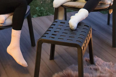 A dark grey, almost black, Nardi Outdoor Resin Balcony Footstool sits on an outdoor wooden deck. Two people are seated on aluminium outdoor chairs nearby, with one person resting their feet on the stool.