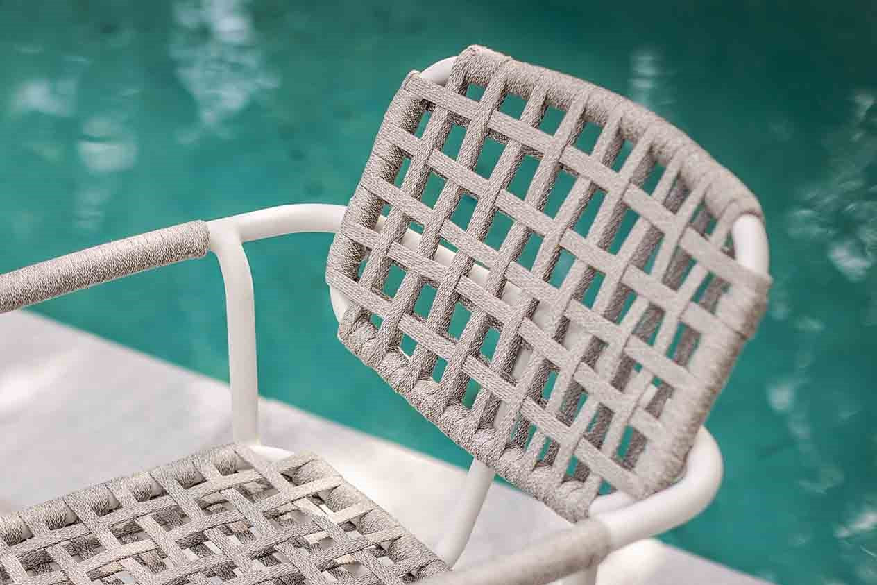 A white outdoor rope chair with a woven seat beside a serene pool reflects a tranquil outdoor setting.