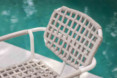 A white outdoor rope chair with a woven seat beside a serene pool reflects a tranquil outdoor setting.