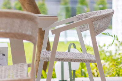 A white table accompanied by two outdoor wicker chairs, nestled in a tranquil garden filled with greenery and blooming plants.