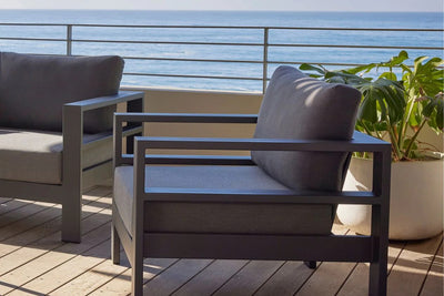 Two sturdy aluminium outdoor armchairs are placed on a wooden deck area overlooking the beach and ocean. Behind one chair, there is a pot plant.