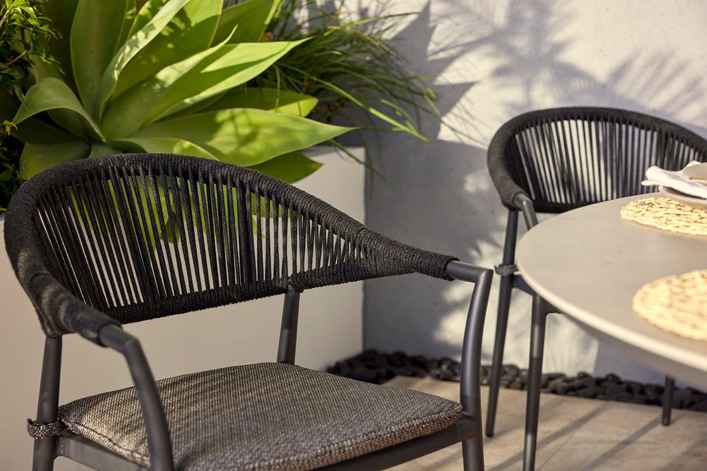 Windsor Rope Outdoor Dining Chairs in Charcoal with grey cushions, featuring a modern rope weave design and sturdy dark metal frame, basking in sunlight in a stylish outdoor space with a light-colored table and lush green plant.
