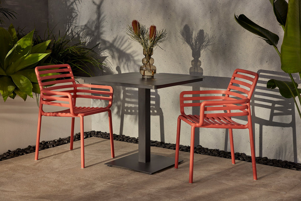 Pair of Nardi Doga Outdoor Resin Dining Armchairs in Marsala positioned on either side of a square black table, adorned with a decorative vase. The ensemble is set against a textured concrete wall featuring surrounding greenery and captivating shadows.