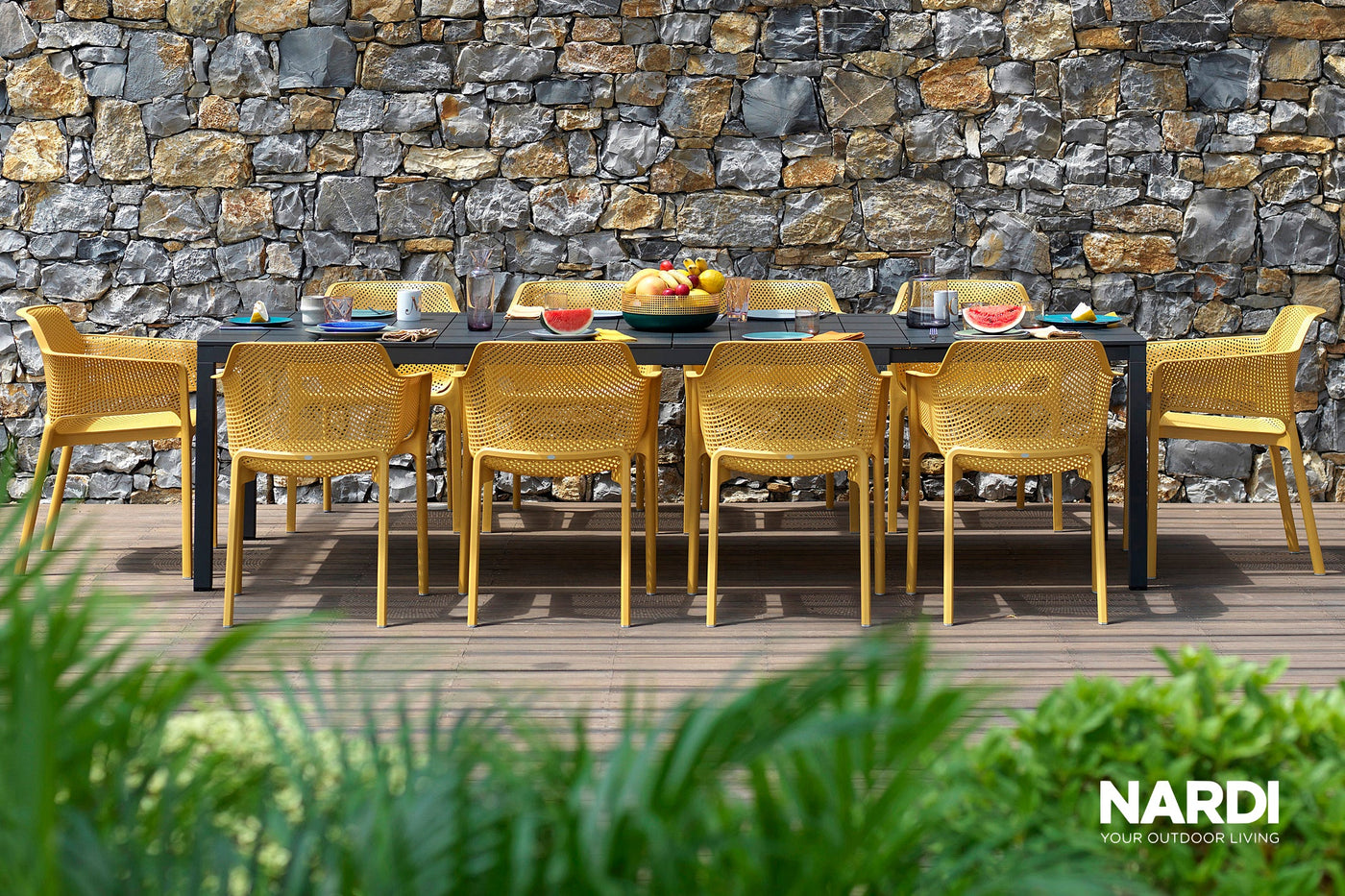 A vibrant outdoor setting featuring some amazing Nardi outdoor furniture pieces. There is a long black extension table and 10 yellow outdoor chairs on a wooden deck, set with food for a welcoming atmosphere.