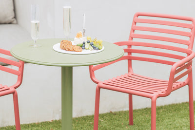 Green outdoor Nardi balcony table set with fruit, crackers, and wine, paired with two salmon pink Nardi chairs in a cozy backyard setting.