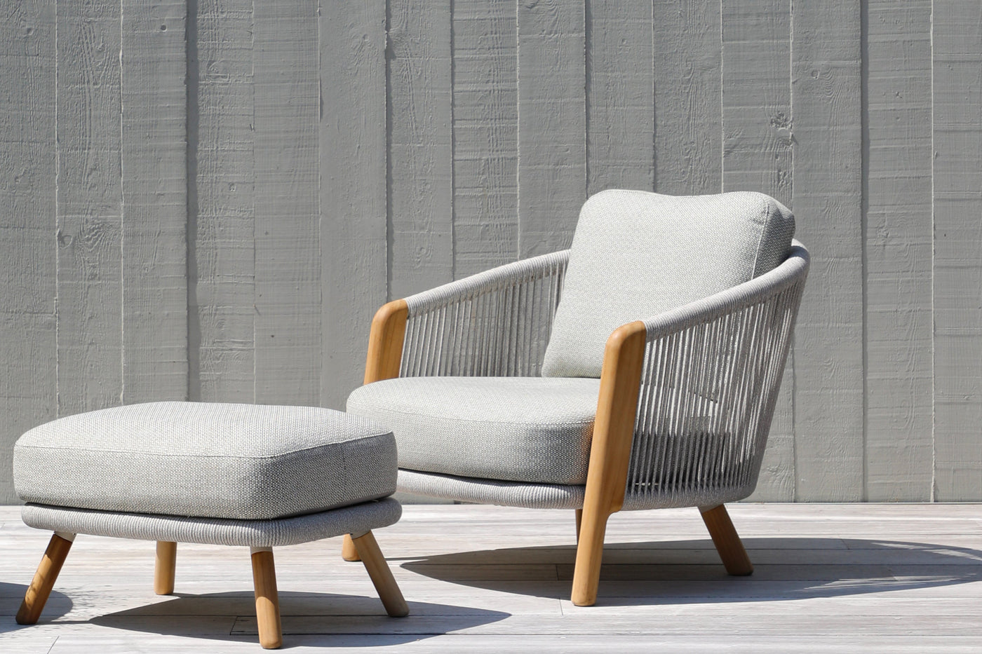 Grey outdoor balcony lounge with matching ottoman on a sunny wooden deck, set against a grey wooden backdrop.