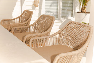 Three tan wicker outdoor bar stools arranged by a table in a cozy backyard, with warm tones of off-white, tan, and light brown for a homey feel.