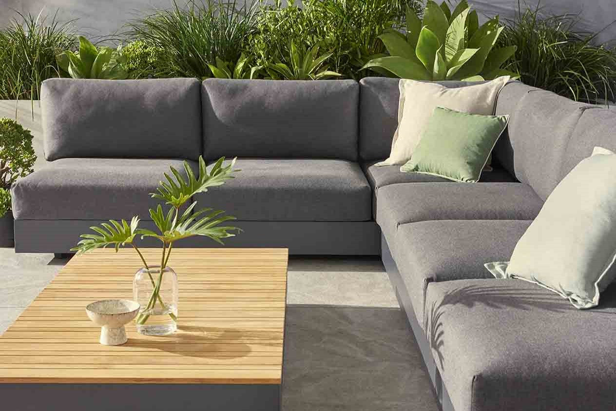 Grey outdoor modular lounge with 3 decorative pillows, set on a patio beside a wooden coffee table. The coffee table features a vase with plants and a decorative small bowl. There is greenery in the background, and the sun is shining on the couch.