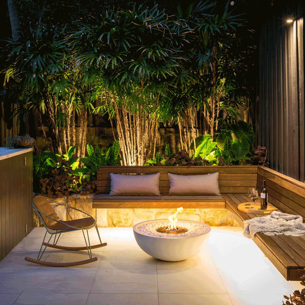 An EcoSmart Mix850 fire pit in bone color is the centerpiece in a cozy outdoor nook with built-in wooden bench seating, soft cushions, a rocking chair, and surrounded by lush tropical plants and ambient lighting.