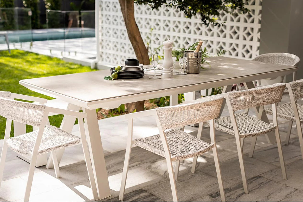 Outdoor dining set featuring five wicker chairs and a ceramic dining table, all with white aluminium frames, placed in a backyard. The table is set with cutlery, and there is greenery behind it. The sun is beaming down, creating an inviting atmosphere.