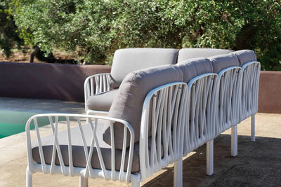 White resin outdoor couch with grey cushions, set by a pool in an outdoor setting, with a large group of trees in the background.