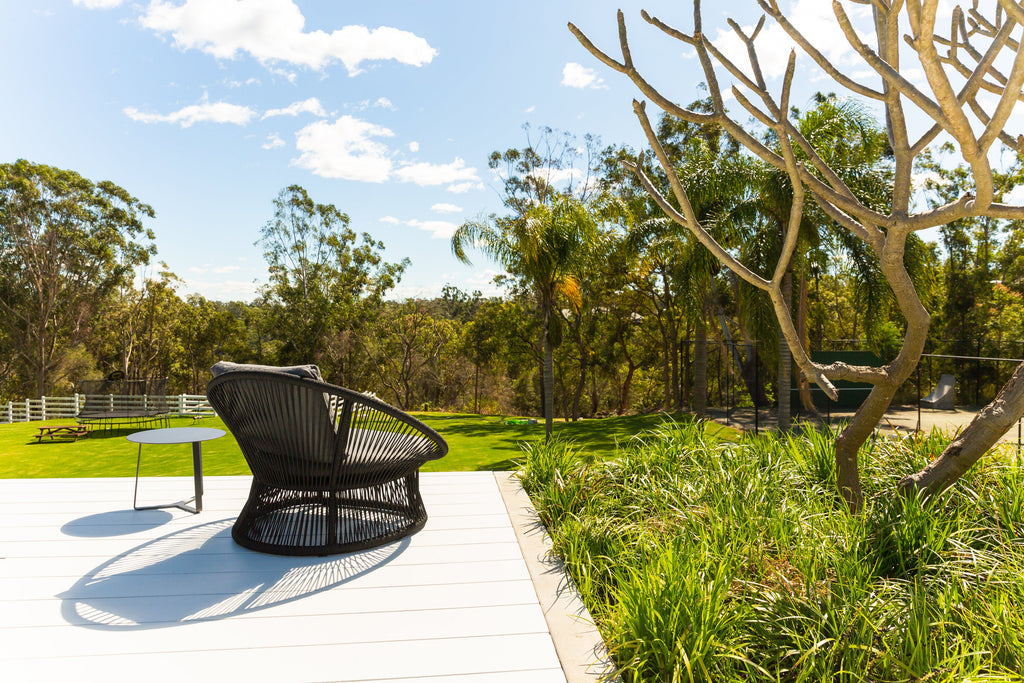 Modern outdoor scene with Spade Wicker Balcony Chair in Black on a white patio, surrounded by greenery, overlooking a serene landscape under a clear blue sky. Sleek Apollo Outdoor Aluminium Round Side Table adds to the contemporary ambiance.