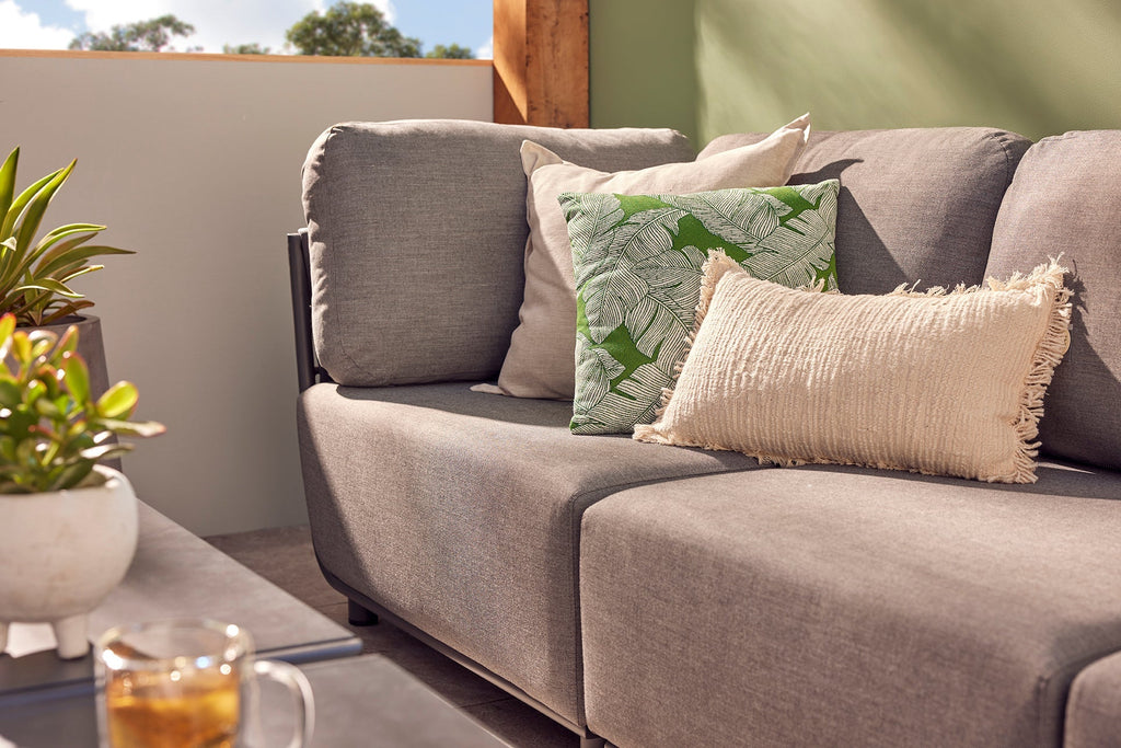 The Lawson Outdoor Lounge in a charcoal grey colour offers comfort with a touch of elegance. It is adorned with decorative pillows, including a vibrant green leaf-patterned pillow and a textured beige fringed pillow basking in warm sunlight.
