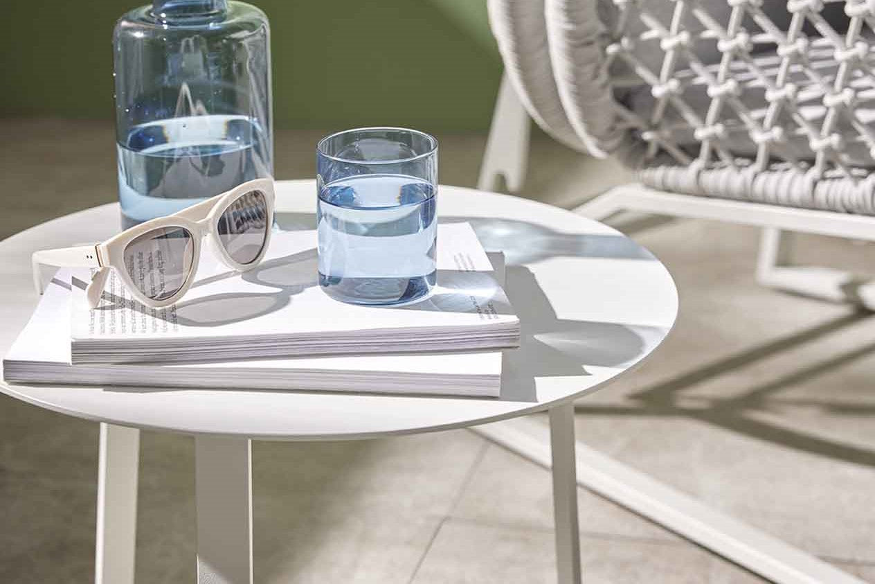 A pair of sunglasses, 2 books, a flask of water, and a glass of water are all set on a round white aluminium outdoor side table with the sun shining in on it. The table is set in a green room with a rope armchair in the background.
