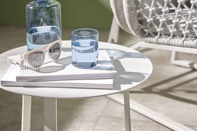 A pair of sunglasses, 2 books, a flask of water, and a glass of water are all set on a round white aluminium outdoor side table with the sun shining in on it. The table is set in a green room with a rope armchair in the background.