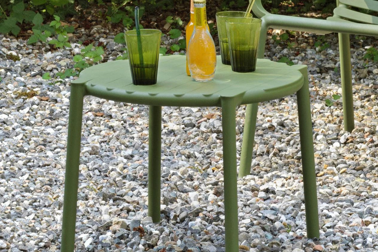 Green resin outdoor coffee table with juice and cups, accompanied by a matching chair, set in a stone area surrounded by lush greenery.