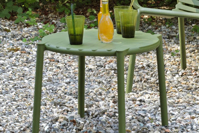 Green resin outdoor coffee table with juice and cups, accompanied by a matching chair, set in a stone area surrounded by lush greenery.