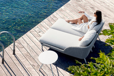 Grey outdoor daybed on a wooden deck beside the ocean, with a woman relaxing in the sunlight.