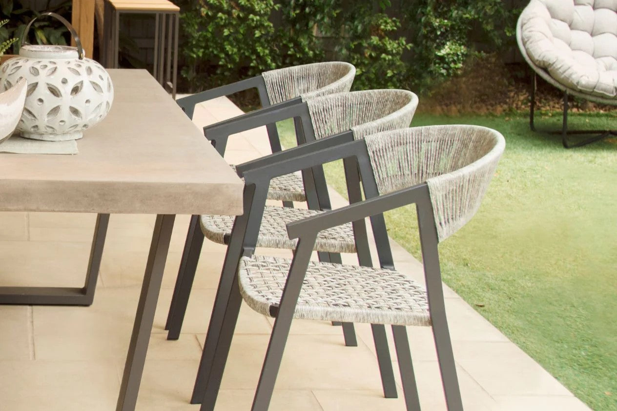Three outdoor wicker dining chairs with charcoal-coloured aluminium frames are facing an outdoor concrete table with a light on it. The setting is in someone's backyard, with greenery visible in the background.